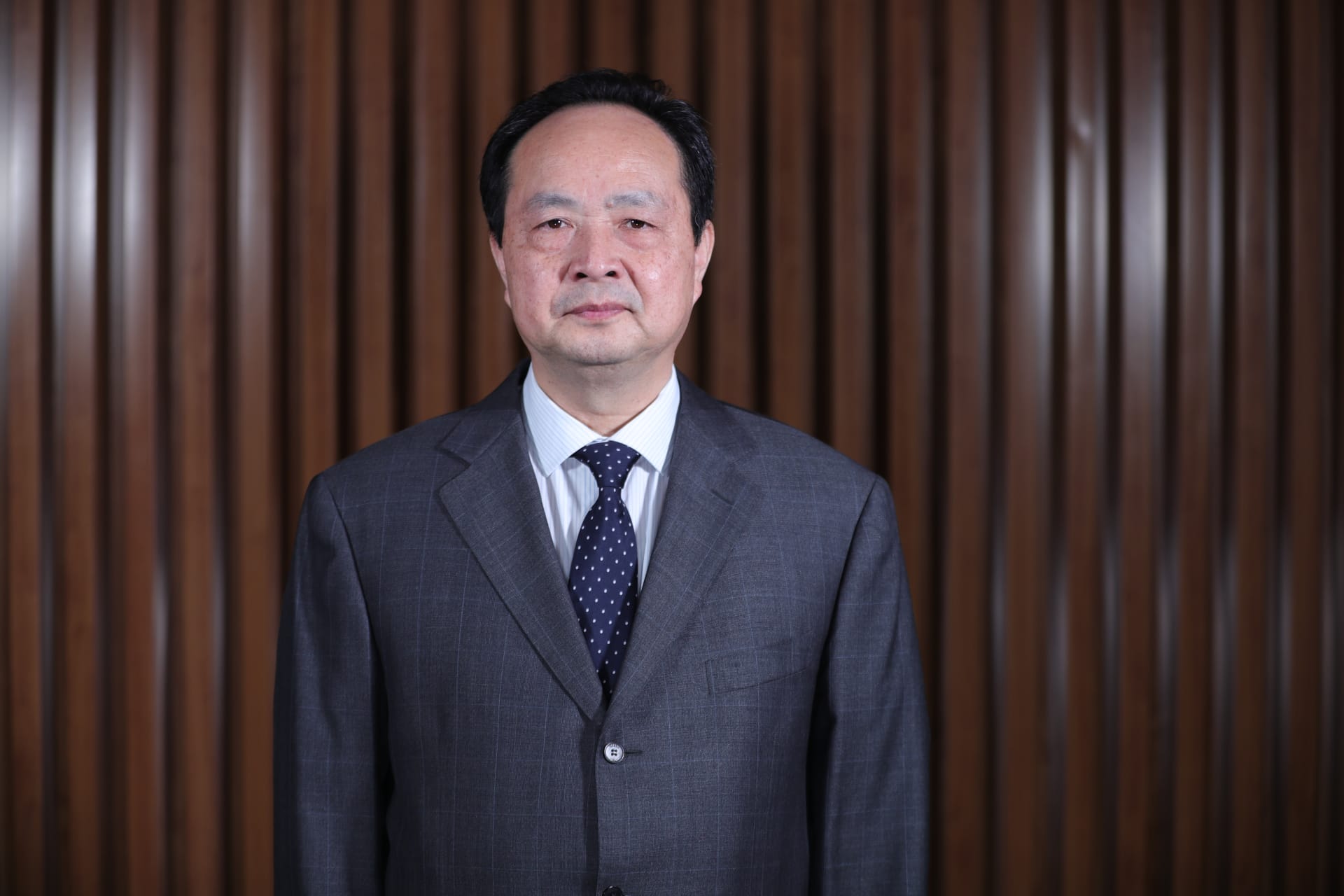 Person in gray suit with blue polka dot tie against wood paneled background.